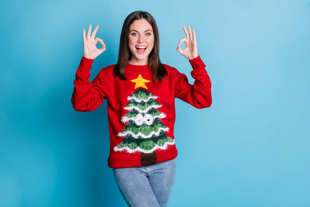 Photo of positive girl in christmas tree pullover sweater show jolly holly okay sign x-mas discounts promo wear denim jeans isolated over blue color background - Zdjęcie, obraz