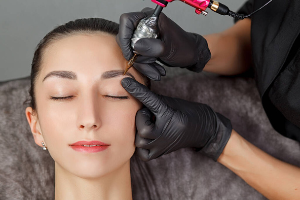 Permanent make up procedure, applying on young girl. Beautiful woman with smooth, young skin, sitting in a beautician. A beautician performs modeling permanent eyebrows makings.Permanent makeup treatment. - Photo, Image