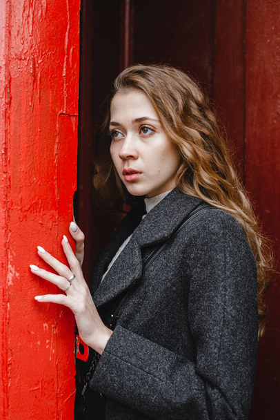 красивая кудрявая девушка стоит возле красной двери в черном пальто - Фото, изображение