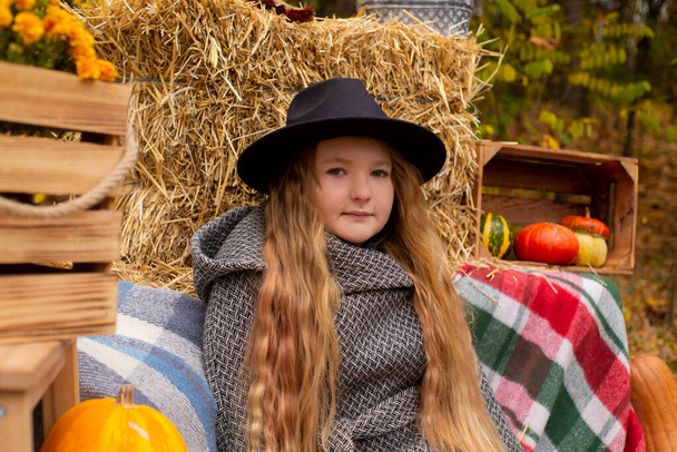 schattig brunette tiener meisje in zwarte hoed en grijze jas in de buurt van de herfst elementen decoratie - pompoenen, appels, plaid, hooi. Gezelligheid, herfst. - Foto, afbeelding