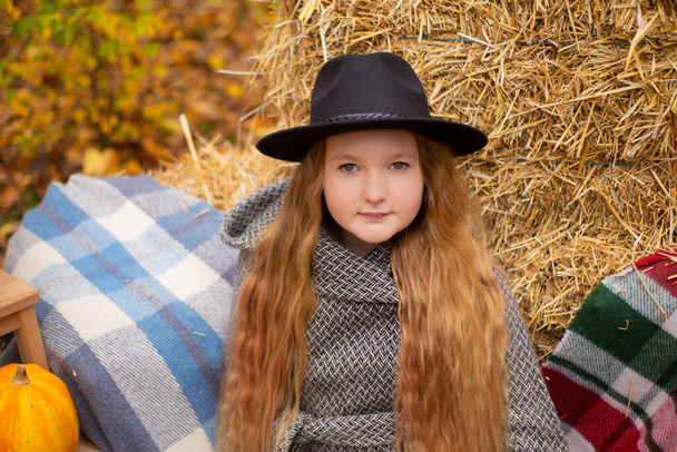 schattig brunette tiener meisje in zwarte hoed en grijze jas in de buurt van de herfst elementen decoratie - pompoenen, plaid, hooi. Gezelligheid, herfst.  - Foto, afbeelding