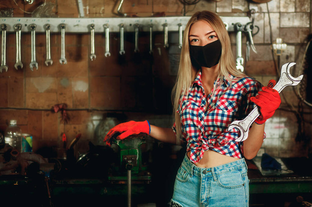 Young girl in denim shorts and a plaid shirt holding a wrench. Girl car mechanic with a mask on her face. Covid-19 pandemic protection concept. Beautiful girl at the service station. - Photo, Image