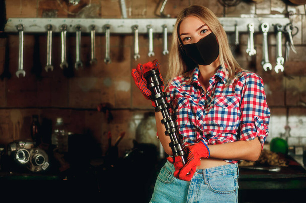 Covid-19 pandemic protection concept. Beautiful girl at the service station. Young girl in denim shorts and plaid shirt holds car engine detail. Engine camshaft in the hands of a beautiful girl - Photo, Image
