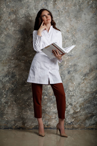 Portret pięknej dziewczyny z długimi falującymi włosami. Kobieta lekarz w białym płaszczu medycznym czytająca z rozmysłem dużą książkę. - Zdjęcie, obraz