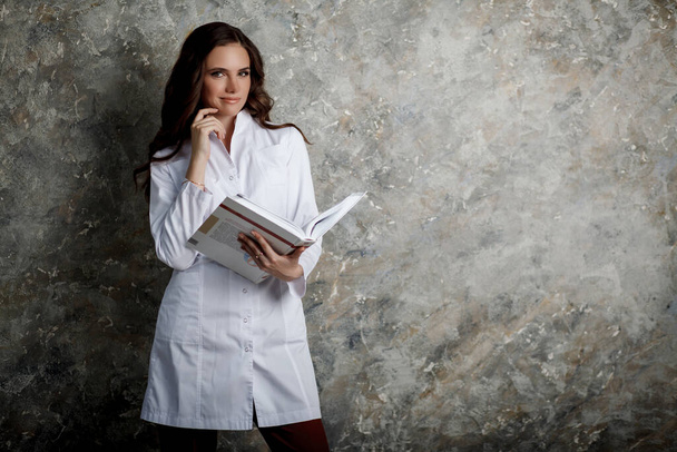Retrato de una hermosa chica con el pelo largo y ondulado. Una doctora con un abrigo médico blanco sostiene un libro grande. Espacio para texto. - Foto, imagen