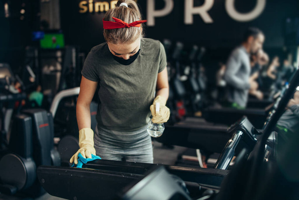 Young female worker disinfecting cleaning and weeping expensive fitness gym equipment with alcohol sprayer and cloth. Coronavirus global world pandemic and health protection safety measures. - Foto, Imagen