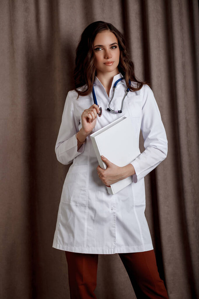 Portrait d'une belle fille aux longs cheveux ondulés. Une femme médecin en manteau médical blanc avec un stéthoscope autour du cou tient un grand livre dans ses mains. - Photo, image