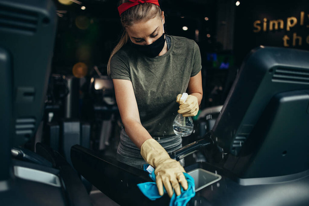Young female worker disinfecting cleaning and weeping expensive fitness gym equipment with alcohol sprayer and cloth. Coronavirus global world pandemic and health protection safety measures. - Fotó, kép