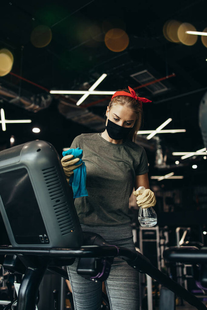 Young female worker disinfecting cleaning and weeping expensive fitness gym equipment with alcohol sprayer and cloth. Coronavirus global world pandemic and health protection safety measures. - Fotó, kép