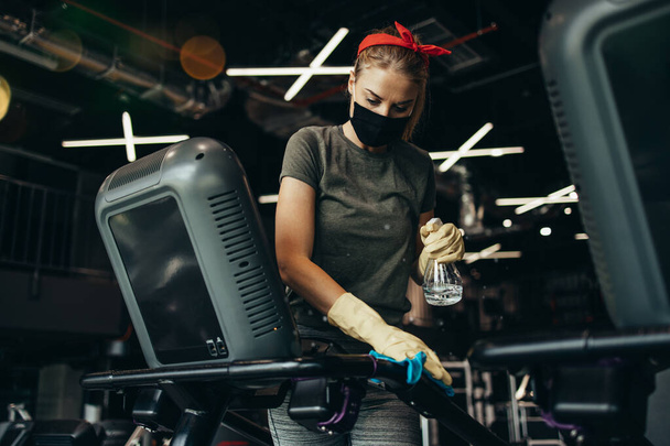 Young female worker disinfecting cleaning and weeping expensive fitness gym equipment with alcohol sprayer and cloth. Coronavirus global world pandemic and health protection safety measures. - Foto, immagini