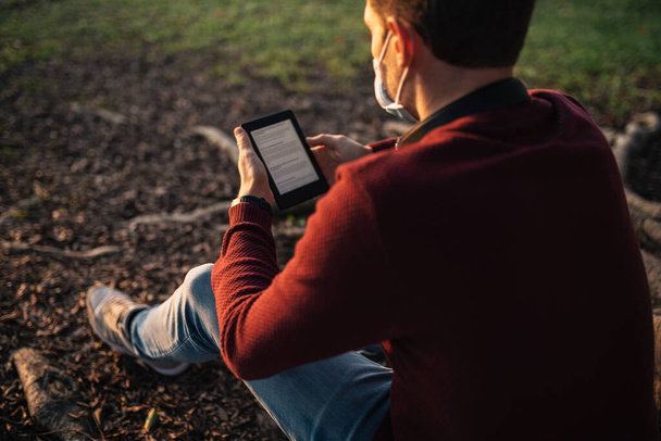 ragazzo con una maschera dal covid-19, leggendo un e-book, seduto su un albero al tramonto. - Foto, immagini