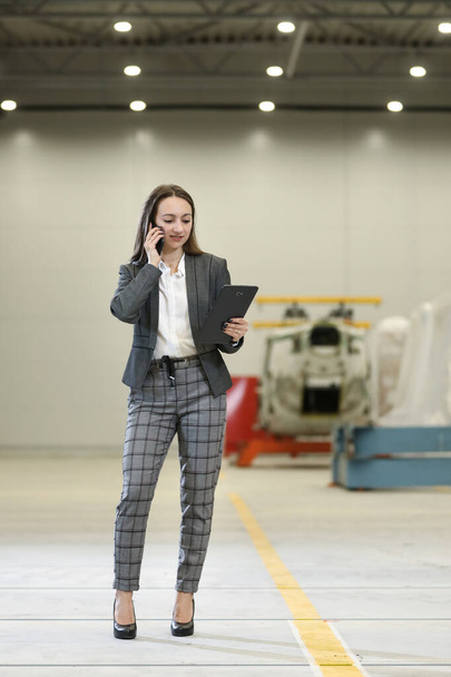 Portré egy női gyár vezetője egy fehér kemény kalap és üzleti öltöny tartó mobiltelefon és tabletta, ellenőrzése a munkafolyamat a helikopter gyártója. - Fotó, kép