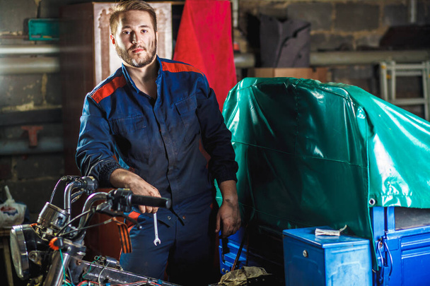 Bir tamircinin portresi. Mavi koruyucu takım elbiseli bir tamirci garajda bir motosikletin yanında duruyor. Araba servisi kavramı. - Fotoğraf, Görsel