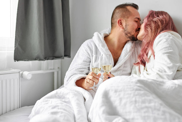 happy lovely couple caucasian man and woman wearing white housecoat, drinking sparkling wine - Foto, imagen
