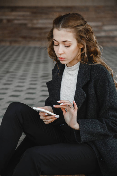 curly-haired girl sitting on street in a black coat and looking at the phone - Photo, Image
