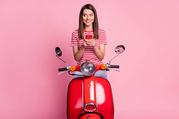 Photo portrait of curious woman sitting on red moped looking at blank space holding cellphone isolated on pastel pink color background - Foto, afbeelding