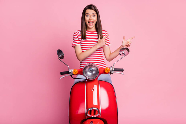 Photo portrait of surprised girl sitting on red moped pointing at blank space smiling cheerfully isolated on pastel pink color background - Foto, afbeelding