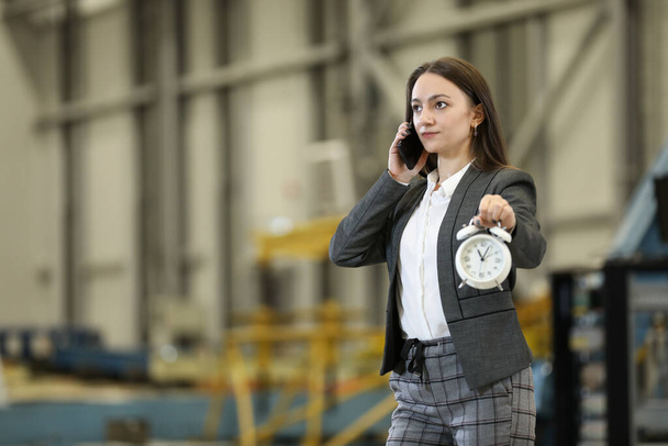 Portrait of a female factory manager in a business suit holding mobile phone and retro alarm clock , controlling the work process in the helicopter manufacturer. Timeline. Deadline. - Zdjęcie, obraz