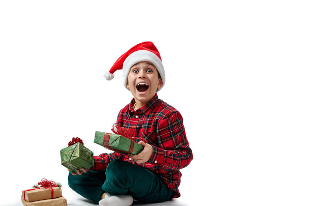 Izolovaný portrét dítěte na bílém. Chlapec v červené košili sedí a raduje se s pusou dokořán, drží vánoční dárky. - Fotografie, Obrázek