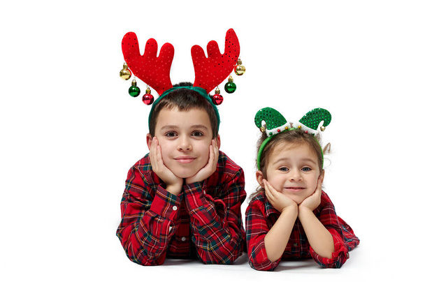 Отдельный портрет на белом. Мальчик и девочка в красных одеждах и рождественских украшениях на их головах, укладывая их лица руками. - Фото, изображение