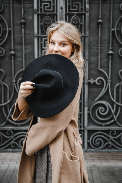 blondýnka v hnědém kabátě s černým kloboukem pózující, usmívající se, rozrušená - Fotografie, Obrázek