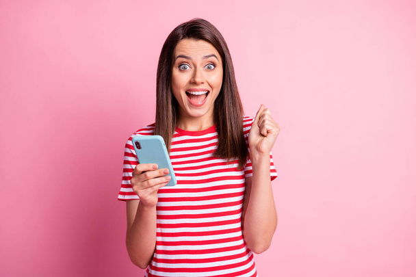 Photo portrait of girl wearing striped t-shirt keeping smartphone gesturing like winner isolated on pastel pink color background - Foto, afbeelding