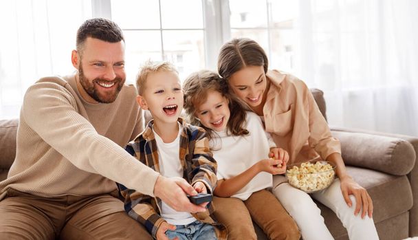 Délicieux parents et enfants de la famille mangeant du pop-corn et riant tout en étant assis sur le canapé et en regardant un film drôle à hom - Photo, image