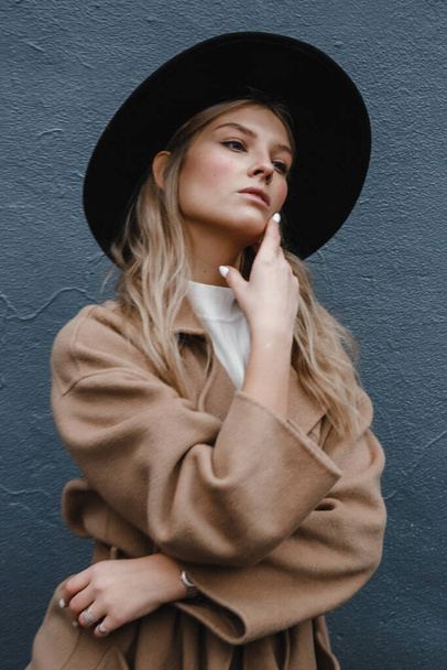 blond in een bruine jas met zwarte hoed poseren, glimlachen, boos - Foto, afbeelding