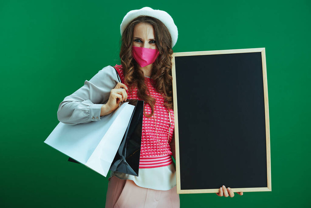 Life during covid-19 pandemic. stylish woman in pink sleeveless shirt with pink medical mask and shopping bags showing blank black board on green background. - Fotoğraf, Görsel