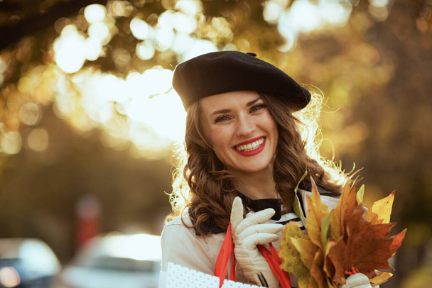 Bonjour septembre. Portrait de femme élégante souriante en trench beige avec sacs à provisions et feuilles jaune automne à l'extérieur dans la ville en automne. - Photo, image