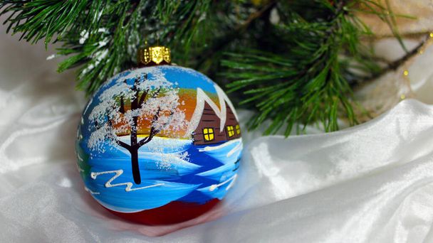 A large glass sphere with a painted winter landscape. Christmas ball hanging on a fir branch on a white silk background. New Year and Christmas ball. - Foto, immagini