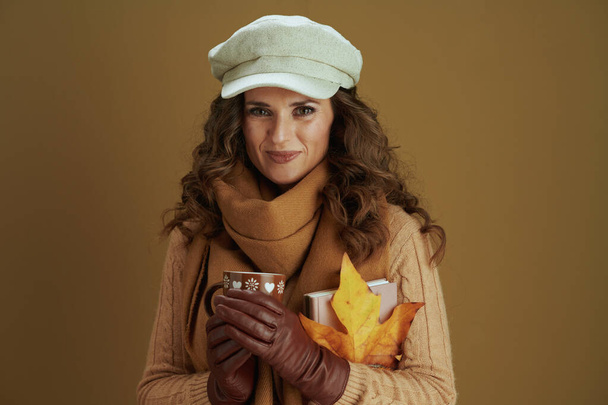 Hello october. Portrait of smiling elegant middle aged housewife in scarf with yellow autumn maple leaf, book, leather gloves and cup against bronze background. - Foto, afbeelding