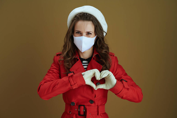 Life during covid-19 pandemic. Portrait of happy stylish 40 years old housewife in red coat showing heart shaped hands isolated on bronze background. - 写真・画像