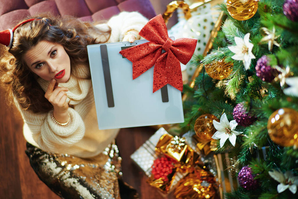 Merry Christmas. Upper view of young female in gold sequin skirt and sweater with scale and red bow near Christmas tree and gift boxes. - Fotografie, Obrázek