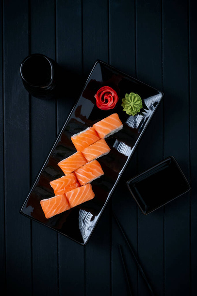Philadelphia-Rollenset mit Lachs, Gurke und Frischkäse auf japanischem Teller auf schwarzem Holzgrund mit Stäbchen und Sojasauce - Foto, Bild