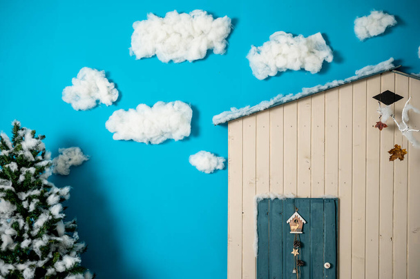Декоративная рождественская студия с домом с голубой дверью, деревянной скамейкой и лыжами, снеговиком, фальшивыми облаками и снегом - Фото, изображение