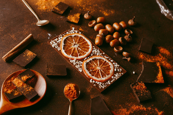 chocolate bar with orange. spices are sprinkled on the surface, orange citron and pieces of dark chocolate on a dark brown background. Topview, copyspace - Zdjęcie, obraz