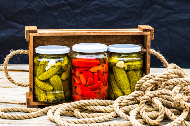Wooden crate with glass jars with pickled red bell peppers and pickled cucumbers (pickles) isolated. Jars with variety of pickled vegetables. Preserved food concept in a rustic composition. - Photo, Image