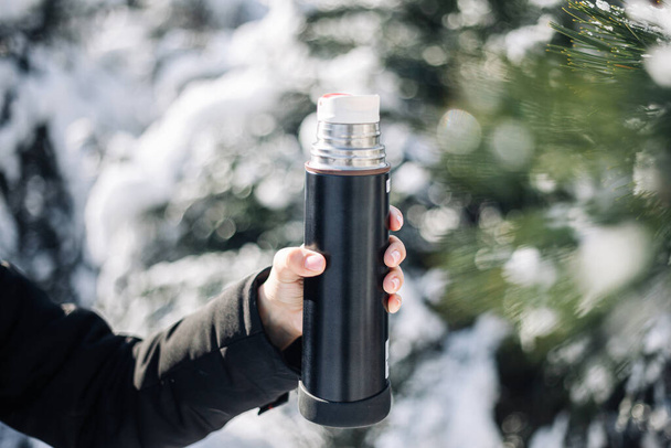 Thermos in the woman's hand on a frosty cold winter day among snowed fir trees in the park. Closeup outdoors shot of a thermos with tea and coffee - Photo, Image
