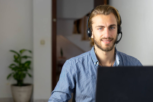 Homme blond travaillant à la maison avec des écouteurs utilisant Internet sans fil sur ordinateur portable, agent du centre d'appels ou télévendeur - Photo, image