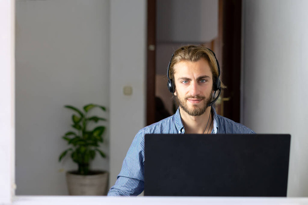 Blond mężczyzna pracuje z domu ze słuchawkami za pomocą bezprzewodowego Internetu na laptopie, człowiek call center agent lub telemarketer - Zdjęcie, obraz