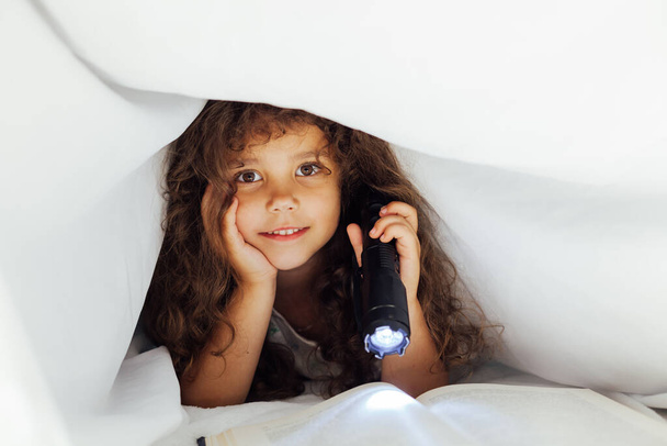 Маленька дівчинка читає книгу вночі з ліхтариком під ковдрою
 - Фото, зображення