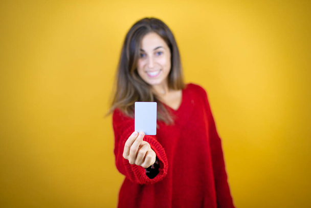 Joven hermosa mujer vistiendo suéter rojo sobre fondo amarillo aislado sonriendo y sosteniendo la tarjeta blanca - Foto, imagen