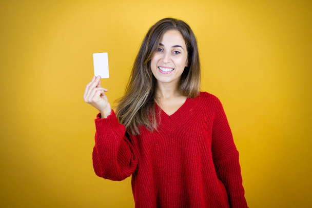 Młoda piękna kobieta w czerwonym swetrze na odizolowanym żółtym tle uśmiechnięta i trzymająca białą kartę - Zdjęcie, obraz