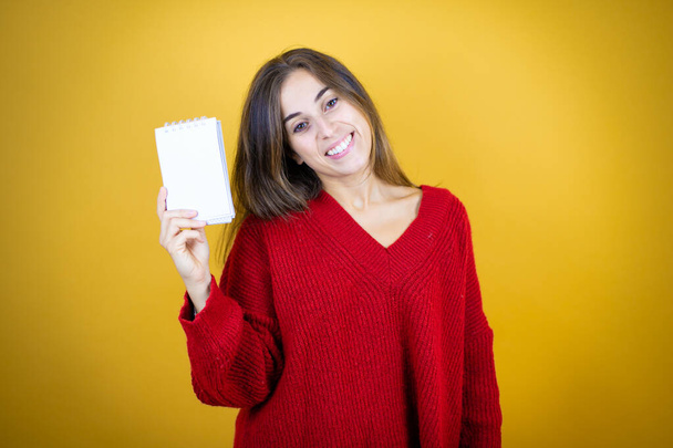 Nuori kaunis nainen yllään punainen pusero yli eristetty keltainen tausta hymyillen ja osoittaa tyhjä muistikirja kädessään - Valokuva, kuva