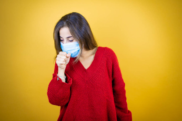 Junge schöne Frau in rotem Pullover und Maske über gelbem Hintergrund mit der Hand vor dem Mund, weil sie hustet - Foto, Bild