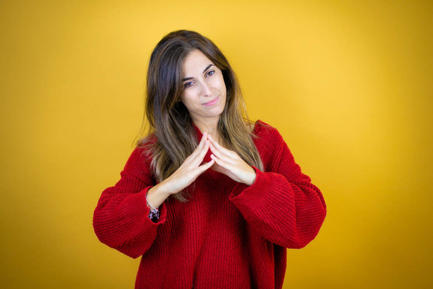 Young beautiful woman wearing red sweater over isolated yellow background with Hands together and fingers crossed smiling relaxed and cheerful. Success and optimistic - Photo, Image