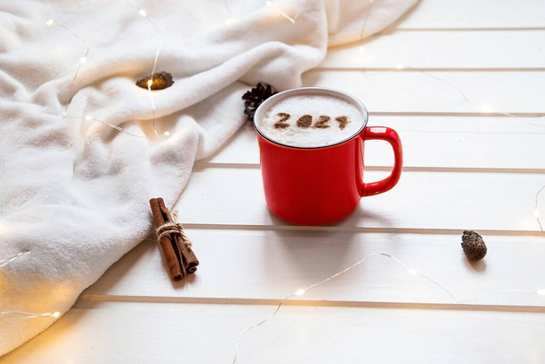 2021-es szám a cappuccino habos felszínén, piros bögrében tálalva a hangulatos kockás és karácsonyi fények hátterén. Élelmiszer művészet kreatív koncepció újév 2021. Lapos, önző fókusz - Fotó, kép