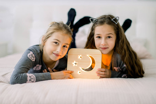 Two pretty little girls sisters, lying on bed at home and enjoying their time playing with wooden night lamp with moon and stars picture. Happy time at home. Cozy home decorations - Zdjęcie, obraz