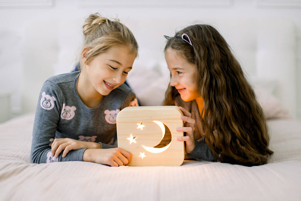Two pretty little girls sisters, lying on bed at home and enjoying their time playing with wooden night lamp with moon and stars picture. Happy time at home. Cozy home decorations - Фото, зображення